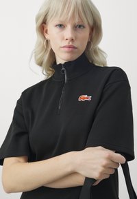 Juodas užtrauktuku užsegamas džemperis su aukštu apykakle, trumpomis rankovėmis, išsiuvinėtu oranžiniu krokodilo logotipu ir lygiu paviršiumi.