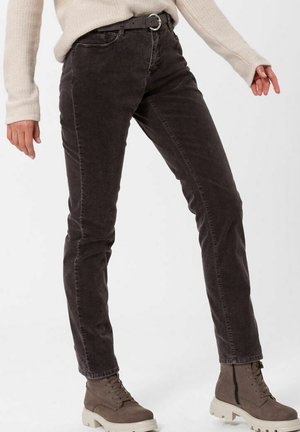 Personne portant un pantalon en velours côtelé marron foncé, un pull en maille beige et des bottines taupe à lacets avec des semelles blanches, debout devant un fond uni.