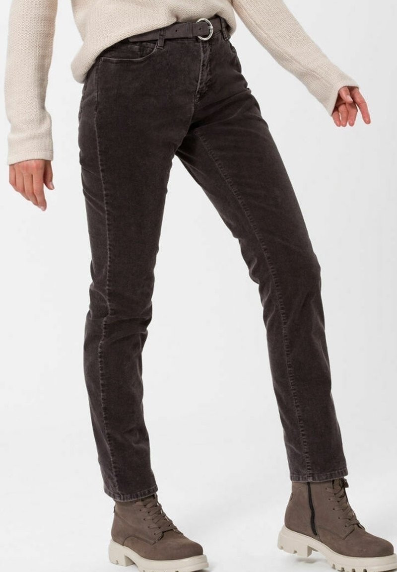 Personne portant un pantalon en velours côtelé marron foncé, un pull en maille beige et des bottines taupe à lacets avec des semelles blanches, debout devant un fond uni.