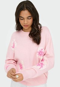 Pull léger rose avec des motifs floraux en rose plus foncé, encolure ras du cou et manches longues. Texture douce, coupe décontractée et poignets côtelés.