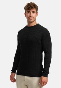Schwarzer Strickpullover mit Zopfmuster, geripptem Rundhalsausschnitt und langen Ärmeln. Über hellen Hosen getragen.