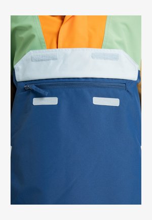 Bunte wasserdichte Jacke mit grünen, orangen und blauen Paneelen. Sie verfügt über eine Reißverschlusstasche mit Klappe und strukturierten Klettverschluss-Akzenten.