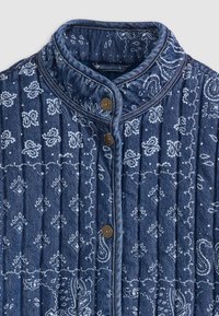 Gequiltede jas in donkerblauwe denim met wit paisleypatroon. Bevat een staande kraag en twee decoratieve messing knopen aan de voorkant.