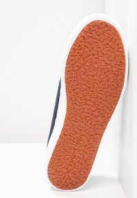 Semelle de chaussure avec bande de roulement en caoutchouc brun texturé et bord blanc, inclinée verticalement sur un fond blanc.