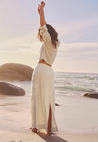 Camicia di lino avorio con maniche a tre quarti e una lunga gonna abbinata con aperture laterali, indossata a piedi nudi su una spiaggia.