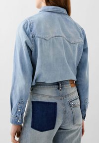 Lichtblauwe denim overhemd met puntkraag en lange mouwen. De achterkant toont een patch zak met donkerblauwe denim op de spijkerbroek, met studaccenten.