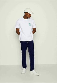 T-shirt branco de manga curta com uma gráfica azul-turquesa no lado esquerdo do peito, combinado com jeans azul-escuro e sapatilhas brancas. O modelo usa um boné claro.
