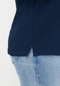 T-shirt blu navy con dettaglio a taglio laterale, realizzato in tessuto testurizzato. Indossato con jeans di denim azzurro, che mostrano una vestibilità rilassata.