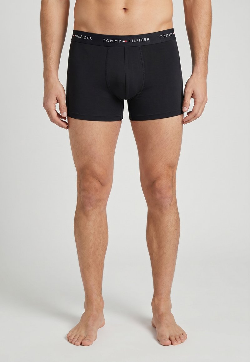 Torse et jambes d’homme portant un boxer noir Tommy Hilfiger avec ceinture marquée, debout pieds nus sur fond uni.