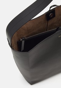 Czarna skórzana torebka o gładkiej powierzchni, z otwartym wnętrzem i brązowym zamszowym wykończeniem. Posiada zasuwaną kieszeń na bezpieczne przechowywanie.