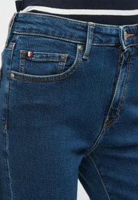 Τζιν από τζιν υλικό σε σκούρο μπλε χρώμα με ελαφρώς φθαρμένη υφή, που διαθέτει πορτοκαλί ραφές, μικρή μπροστινή τσέπη και ετικέτα με λογότυπο.