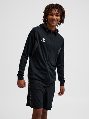 Lächelnder junger Mann mit Dreadlocks, der einen schwarzen Kapuzenpullover mit Reißverschluss und schwarze Sportshorts trägt, steht vor einem einfarbigen hellen Hintergrund.