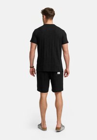 Schwarzes Kurzarmhemd und Shorts-Set aus strukturiertem Stoff. Mit einem kleinen Etikett auf der Rückseite der Shorts. Tragen mit Schlupfsandalen.