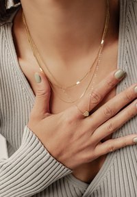 Goldene, geschichtete Halsketten mit einer strukturierten Kette, flachen Gliedern und einem kleinen Herzanhänger, getragen über einem gerippten grauen Oberteil. Hände mit grüner Nagellackfarbe.
