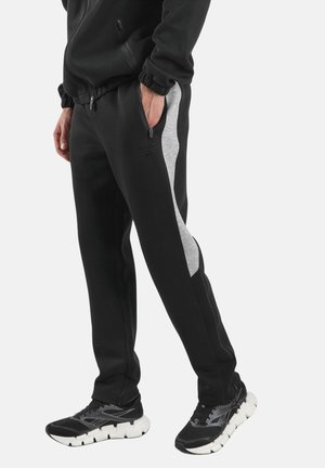 Man draagt zwarte trainingsbroek met grijze zijpanelen en zwarte sportschoenen met witte zolen, staat met handen in zakken.