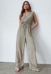 Combinaison sans manches beige clair avec un devant à boutons, taille élastique, poches latérales et jambes larges, fabriquée dans un tissu léger et texturé.
