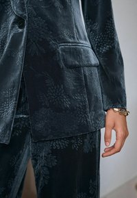 Sacou catifelat bleumarin, cu un model floral embosat, având un singur nasture la închidere și buzunar frontal. Accentuat cu o brățară aurie.