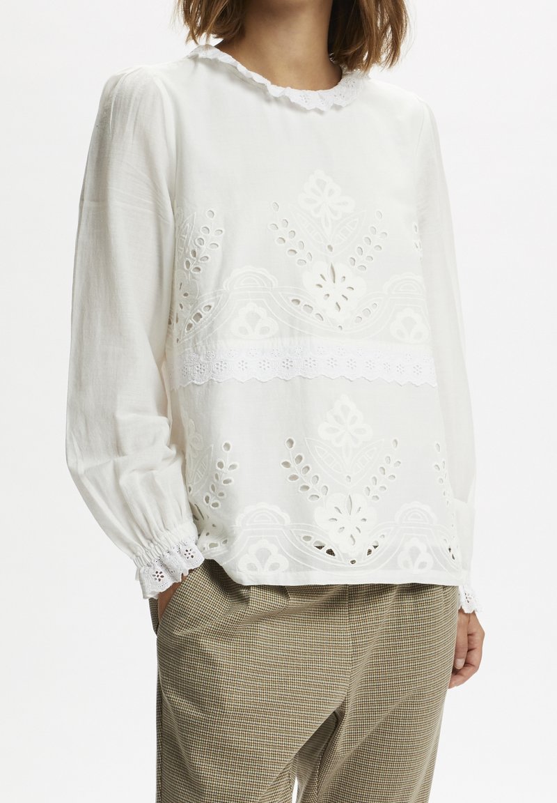 Femme portant un chemisier blanc à manches longues avec broderie florale ajourée et bords festonnés, associé à un pantalon à carreaux marron.