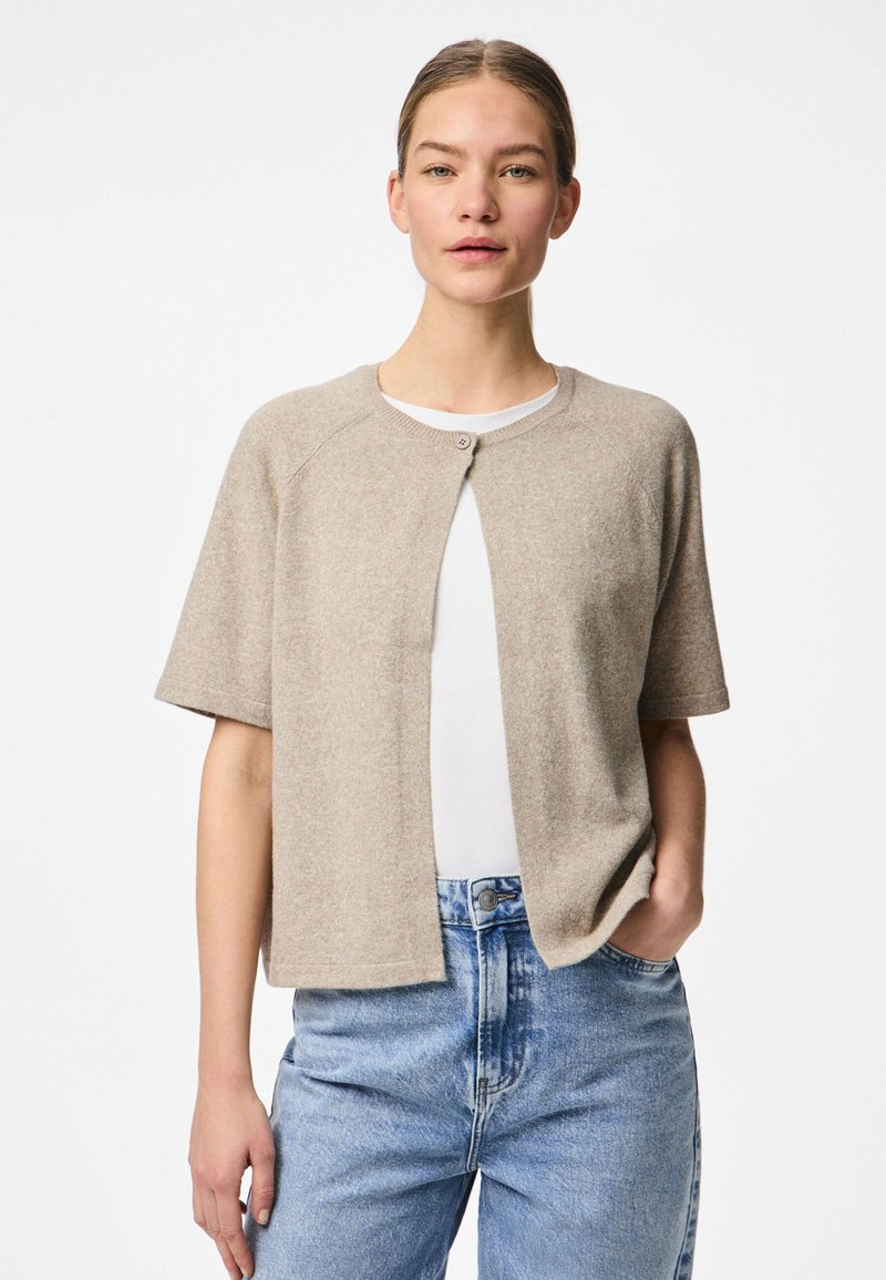 Cardigã bege de mangas curtas com decote redondo e fecho de botões, feito de um material suave, combinado com jeans de denim azul-claro.