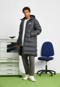 Crna puffer jakna s kapuljačom, zatvaračem i bijelim Nike logom. Uparena s sivim hlačama i bijelim tenisicama. U pozadini se nalazi kopirka i plava stolica.