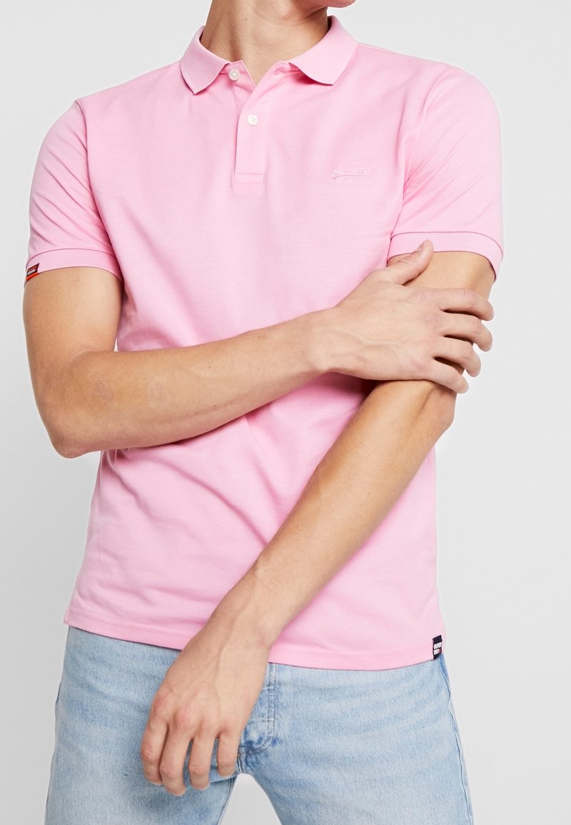 Lyserød polo-skjorte med et klassisk design, korte ærmer og krave med to knapper. Har broderet logo og ribbede manchetter. Parret med lyseblå jeans.