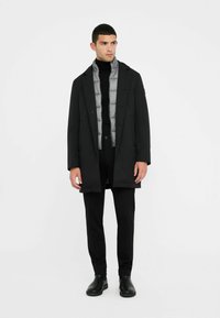 Schwarzer Obermantel über einer grauen Steppweste, schwarzer Rollkragenpullover und maßgeschneiderte Hose. Schwarze Ankle Boots. Minimalistisches Design mit klaren Linien.