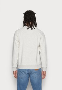 Persoon met schouderlange dreadlocks staat met de rug naar de camera, draagt een lichtgrijze sweatshirt en blauwe spijkerbroek met zichtbaar achterzakken.