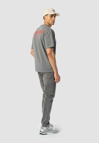 Camiseta de algodón gris con estampado de letras rojas en la parte posterior, combinada con jeans grises y zapatillas de deporte plateadas; gorra beige en la cabeza del modelo.