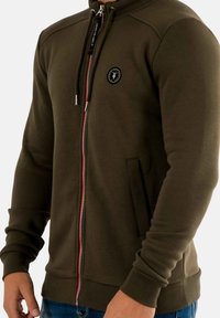 Olijfgroen zip-up hoodie met een hoge kraag, voorzien van een rode rits, zijzakken, geribbelde manchetten en een ronde logo patch op de borst.