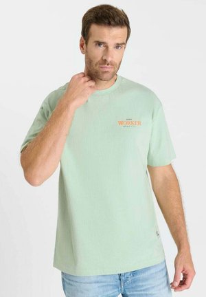 Man met kort bruin haar, gekleed in een lichtgroen T-shirt met oranje tekst "WERKNEMER", staand tegen een effen witte achtergrond.