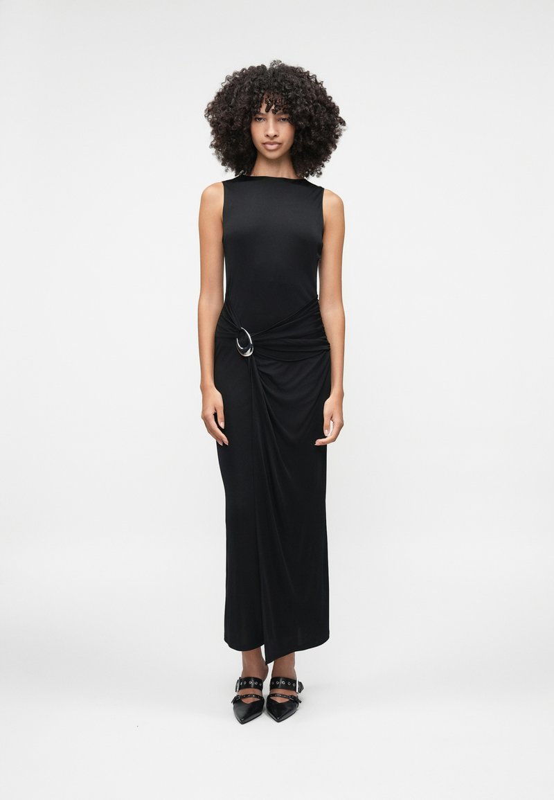 Μαύρο αμάνικο μακρύ φόρεμα με στριμμένη λεπτομέρεια στη μέση, διακοσμημένο με έναν μεγάλο ασημί δακτύλιο. Κατασκευασμένο από λεία ύφανση.