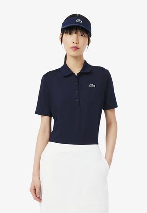 Femme portant un polo bleu marine et une visière assortie avec un logo de crocodile vert, associée à une jupe blanche, debout devant un fond blanc.