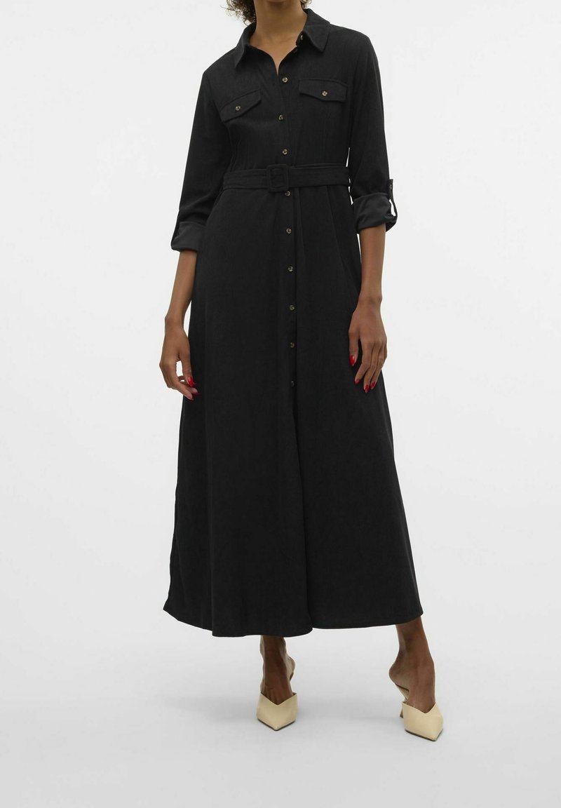 Femme portant une longue robe noire boutonnée avec ceinture, manches remontées, et des escarpins beiges à bout pointu, debout devant un fond uni.