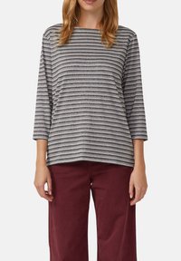 Vrouw in een grijs en zwart horizontaal gestreept lange mouwen top, gecombineerd met losse donkerrode broeken, staande tegen een effen achtergrond.