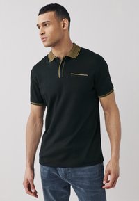 Juodas trumpomis rankovėmis polo marškinėlis su juostuotu aukštu kaklu, puoštas auksinėmis detalėmis, su sagų juosta ir krūtinės kišene. Minkšta medžiaga, aptempiančio silueto dizainas.