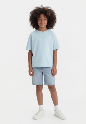 Ung person med krøllet hår iført en lyseblå t-shirt, denimshorts, hvide sokker og hvide sneakers, stående og smilende.