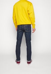 Sudadera amarilla, vaqueros oscuros y zapatillas rojas. La sudadera tiene un cuello redondo; los vaqueros presentan un diseño en el bolsillo trasero con un logotipo.