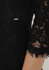 Robe en dentelle noire présentant des motifs floraux complexes, des bords de manches effilochés, et une petite étiquette métallique avec l'inscription "NORMANI" attachée.