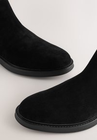 Melni zamšādas potīšu zābaki ar apaļiem purngaliem, minimālu šuvju detalizējumu un kontrastējošu melnu zoli. Virsmas tekstūra ir gluda.