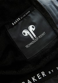 Étiquette en tissu noir avec une icône d'écouteurs blancs et le texte "BAKER BY TED BAKER" et "POCHETTE TECHNOLOGIE" brodé sur un matériau en polaire noir.
