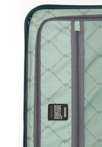 Interior de tela verde de una maleta con un patrón repetitivo de "WITTCHEN", cremallera gris, bolsillo de malla y una etiqueta para la identificación de la marca.