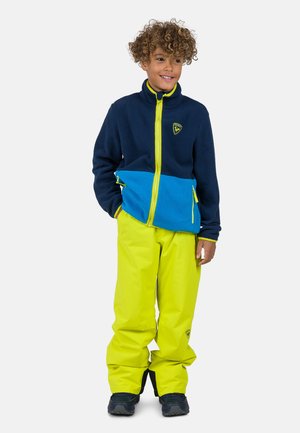 Giacca in pile blu e navy con una cerniera gialla, abbinata a pantaloni impermeabili giallo brillante. Presenta tasche con cerniera e un tessuto testurizzato.