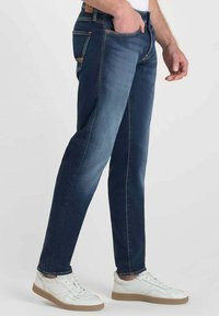 Jeans de mezclilla azul oscuro con un diseño ajustado, que cuentan con cinco bolsillos y un ligero desgaste en los muslos. Combinados con zapatillas blancas.