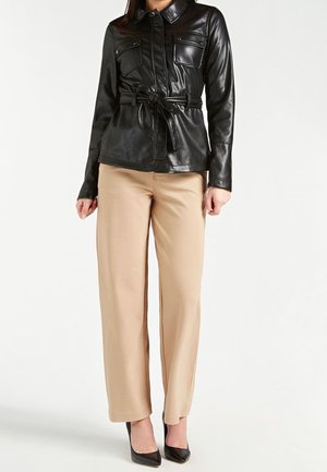 Mujer con chaqueta negra de cuero sintético con cinturón, pantalones beige de pierna ancha y tacones negros puntiagudos, de pie contra un fondo blanco.