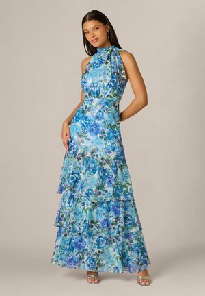 Kobieta ubrana w niebieską kwiecistą, warstwową suknię maxi bez rękawów z kokardką przy wysokim dekolcie, pozująca z jedną ręką na biodrze na jednolitym tle.