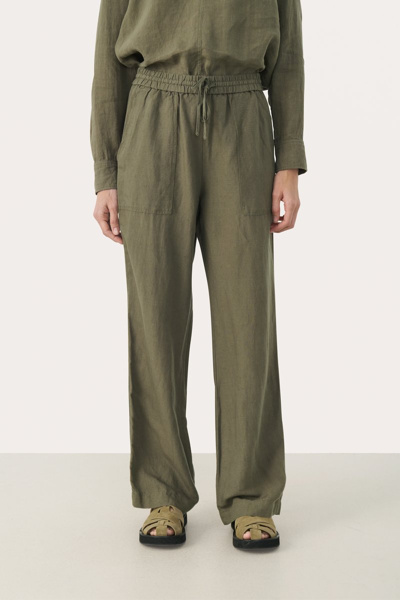 Olijfgroene linnen broek met een elastische tailleband, twee voorzakken en een losse pasvorm, gecombineerd met tan kleurige strappy sandalen.