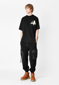 Camiseta negra oversized con diseño gráfico en el lado izquierdo del pecho, combinada con pantalones cargo negros que tienen grandes bolsillos laterales y botas naranjas.