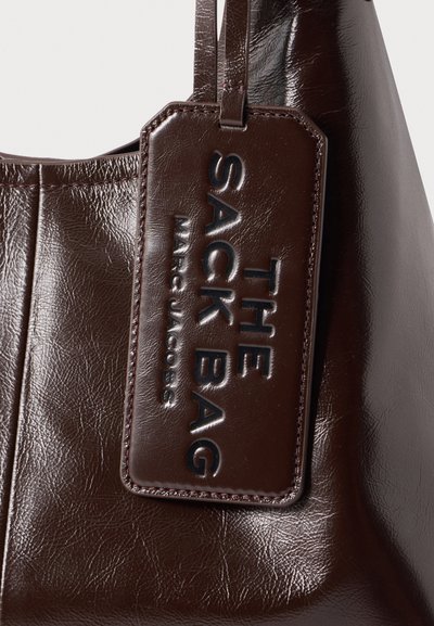 Тъмнокафява кожена чанта с подходящ кожен етикет с дебосван надпис "THE SACK BAG MARC JACOBS", прикрепен към дръжката ѝ.