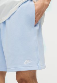 Κοντινό πλάνο ενός ατόμου που φοράει ανοιχτό μπλε σορτς Nike με το χέρι στην τσέπη, εμφανίζοντας το άσπρο λογότυπο Nike κοντά στο τελείωμα.