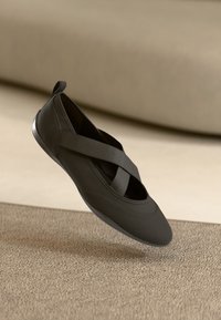 Sapatilha preta de encaixe com tiras elásticas cruzadas e bico arredondado, posicionada acima de um tapete bege texturizado e fundo claro.